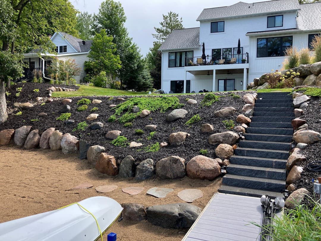 Landscaped hillside with stone steps leading to a modern white house by a lake.