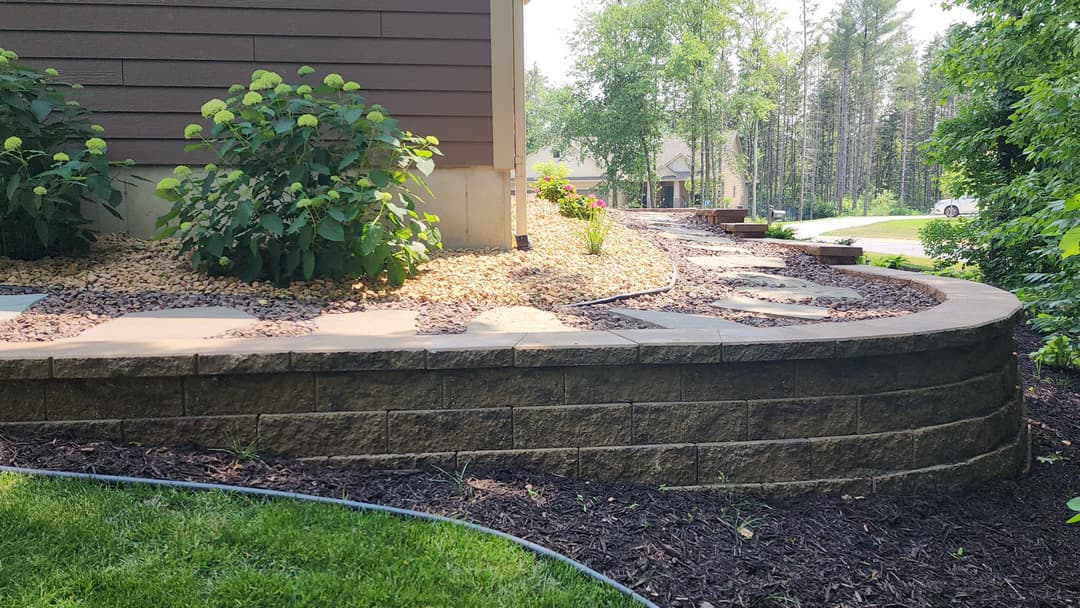 Landscape design featuring a curved stone wall, gravel path, and lush greenery by a house.