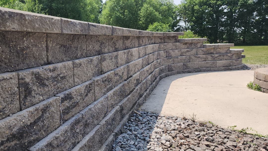 Curved stone retaining wall with textured blocks, surrounded by greenery and gravel path.