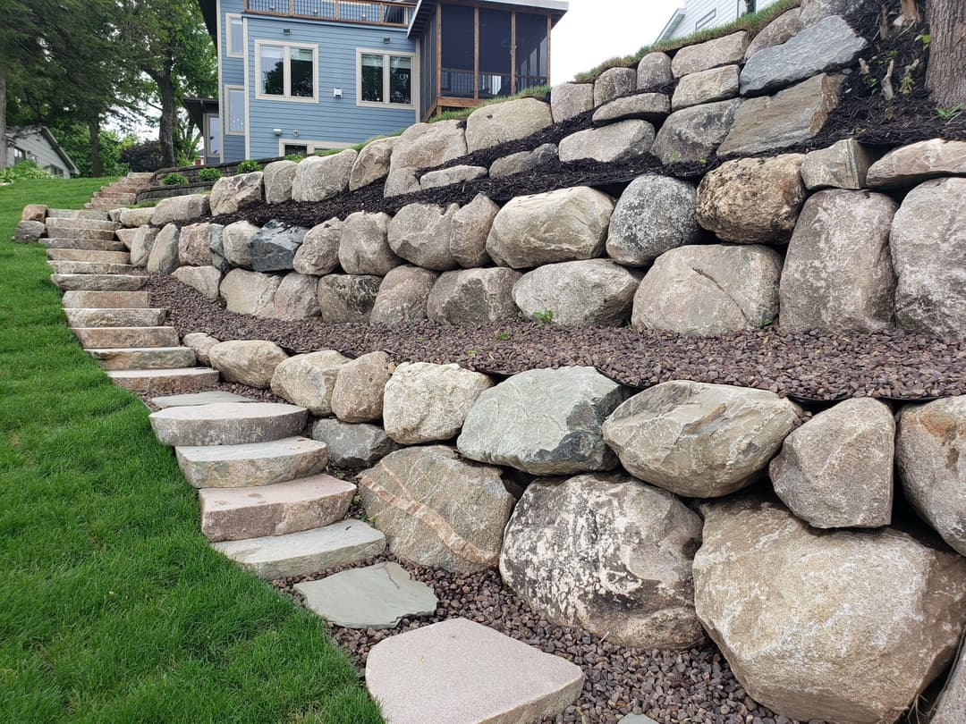Stone retaining wall with steps leading to a house, surrounded by green grass and landscaping.