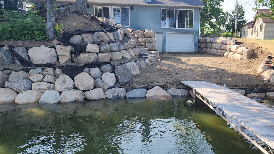 Rock retaining wall by a lake with a wooden dock and a house in the background.