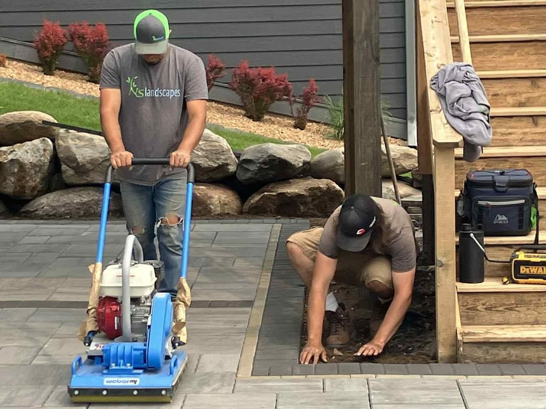 Two landscapers working on a patio installation, one using a compactor and the other leveling stones.