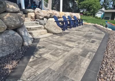 Patio area with stone steps, large rocks, and blue Adirondack chairs near a grassy yard.