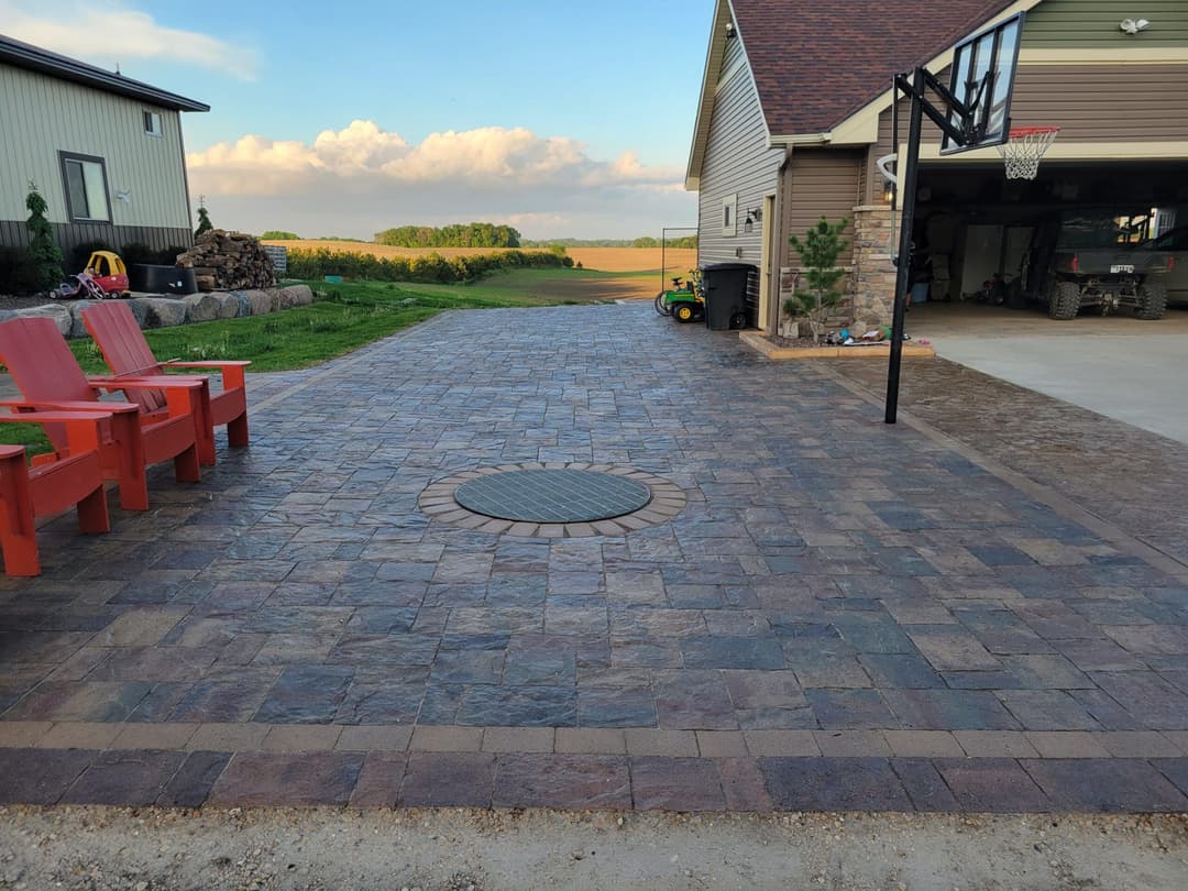 Paved driveway with circular drain cover, red chairs, and a home in a rural setting.