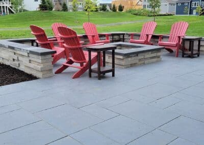 Outdoor patio with red Adirondack chairs around a stone fire pit, surrounded by green lawn.