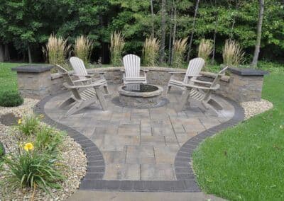 Outdoor fire pit area with stone seating and landscaping, featuring four Adirondack chairs.