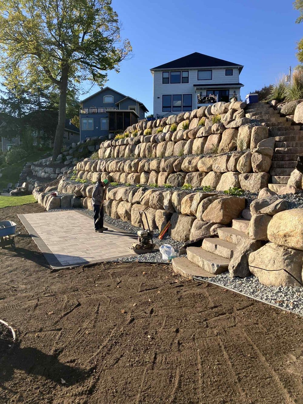 Landscaper working on stone retaining wall and patio installation in residential yard.
